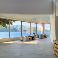 Amplia habitación con grandes ventanales que ofrecen vista a un lago y montañas. Hay varios sofás y una mesa en la habitación.