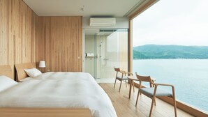 A modern hotel room with a large bed, a seating area with chairs, a glass shower, and a view of a body of water.