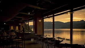 Vista interna di un ristorante con area bar, tavoli e sedie, affacciata su un corpo d'acqua con montagne sullo sfondo.