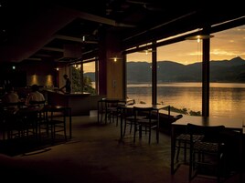 Vista interior de un restaurante con barra, mesas y sillas, con vistas a una masa de agua y montañas al fondo.