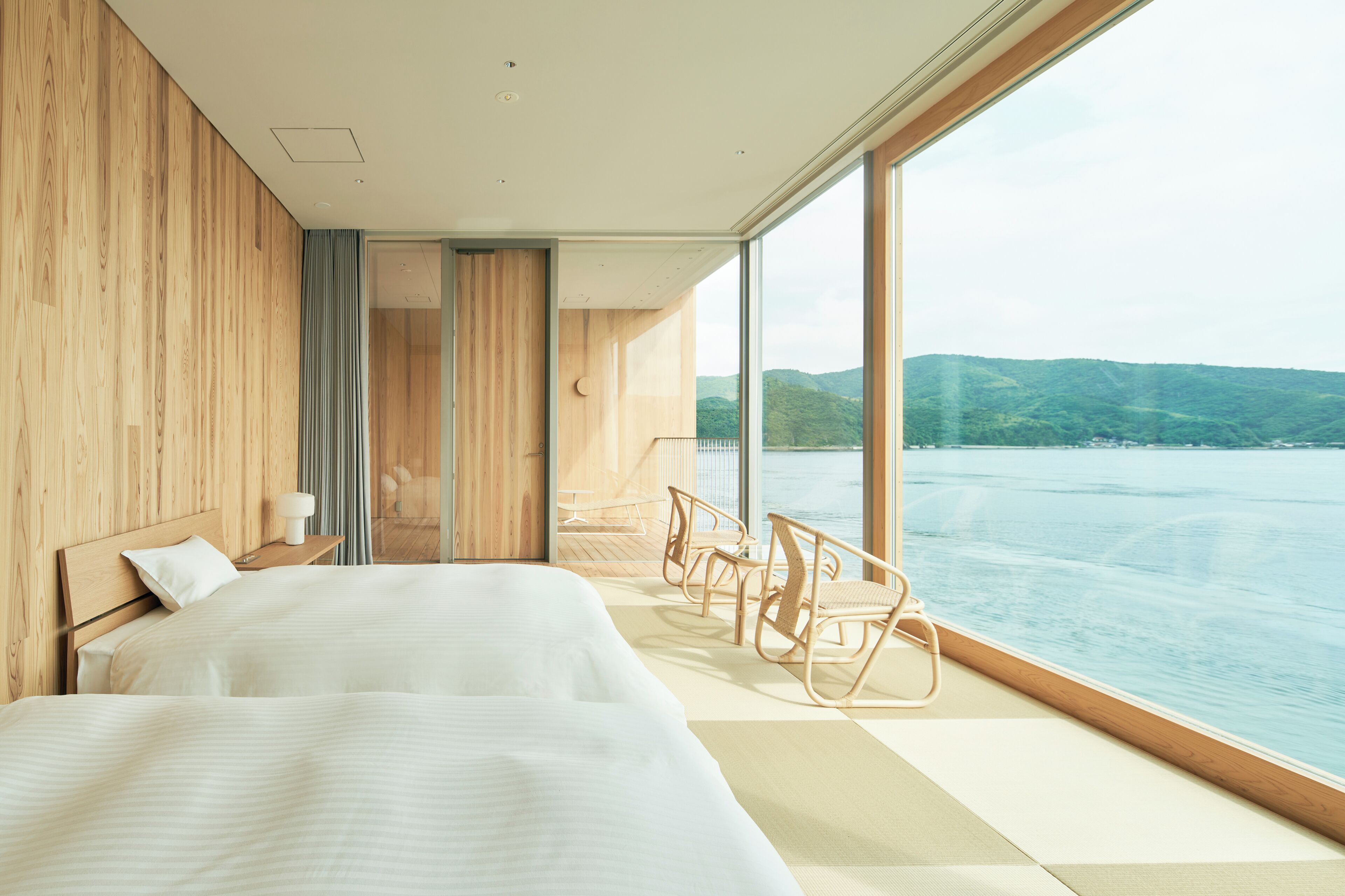 A modern hotel room with a large window, two beds, wooden paneling, and a view of a body of water.