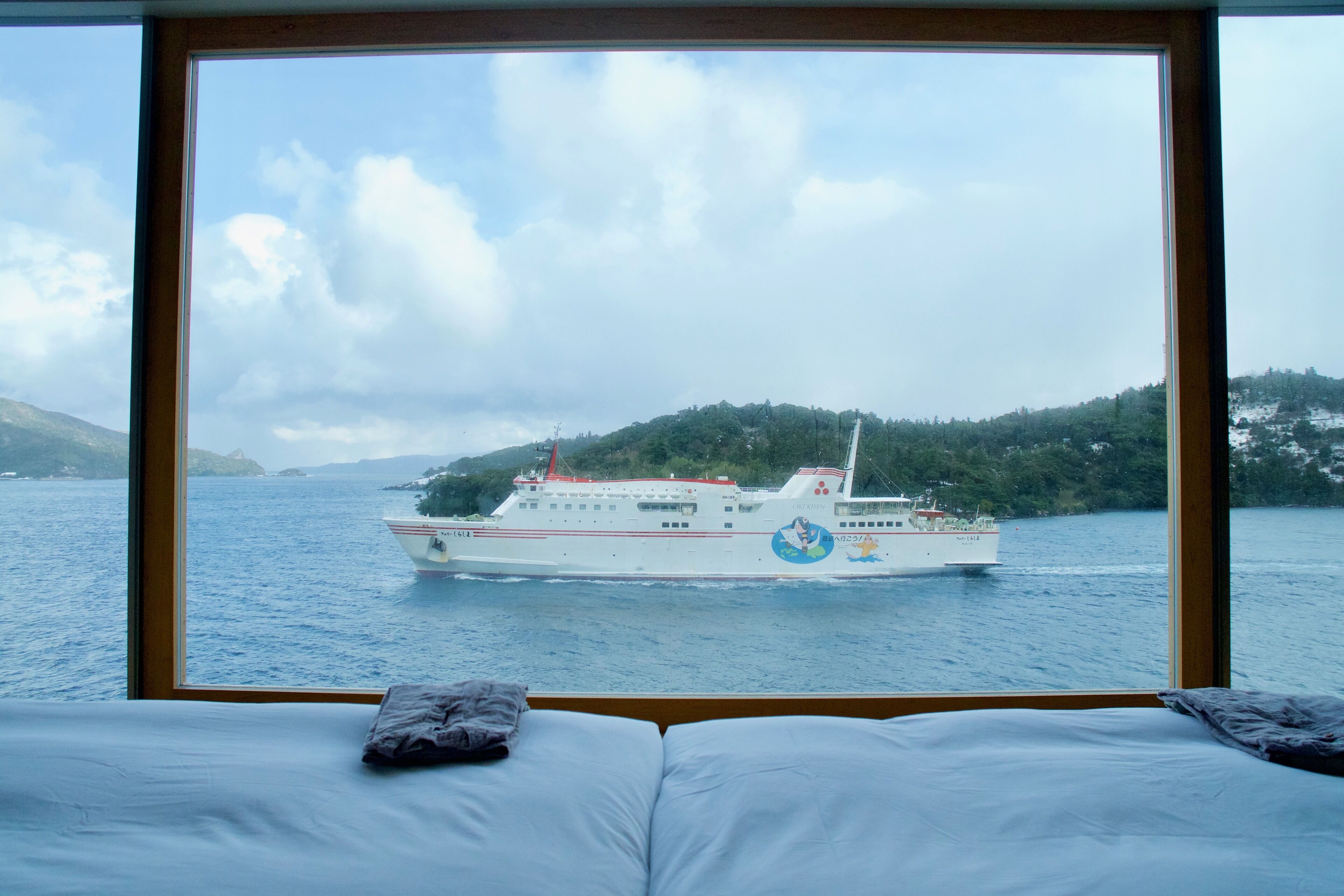 A bed with a view of a ferry boat on a body of water, surrounded by greenery.