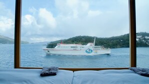 A bed with a view of a ferry boat on a body of water, surrounded by greenery.