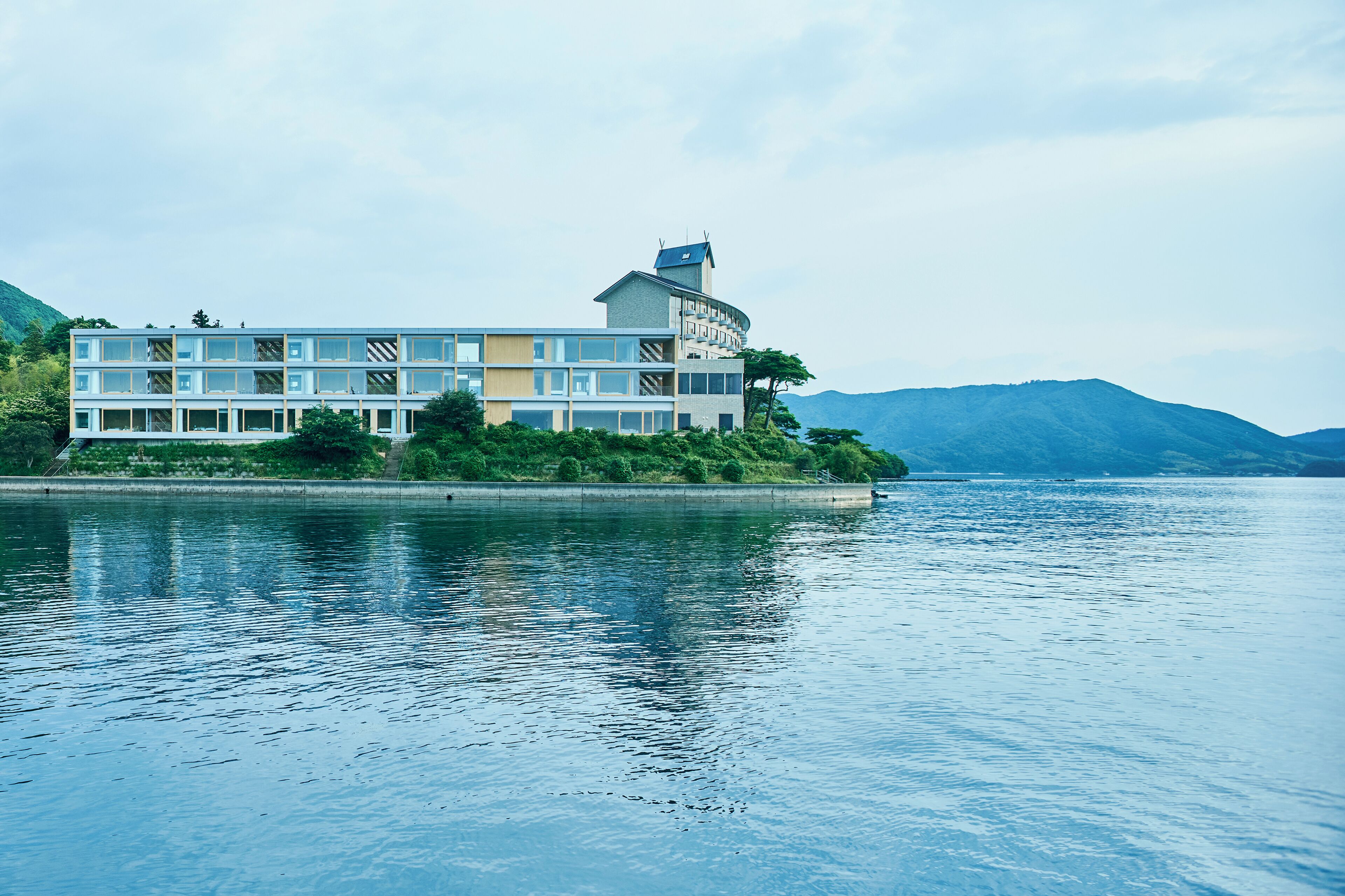A modern hotel with multiple floors, a rooftop terrace, and a view of a lake and mountains.
