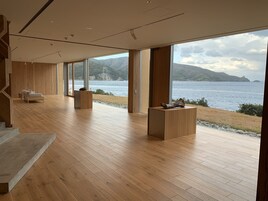 Una habitación amplia con suelo de madera, ventanas grandes que ofrecen vistas a un lago y montañas, un sofá y dos mesitas de noche de madera.