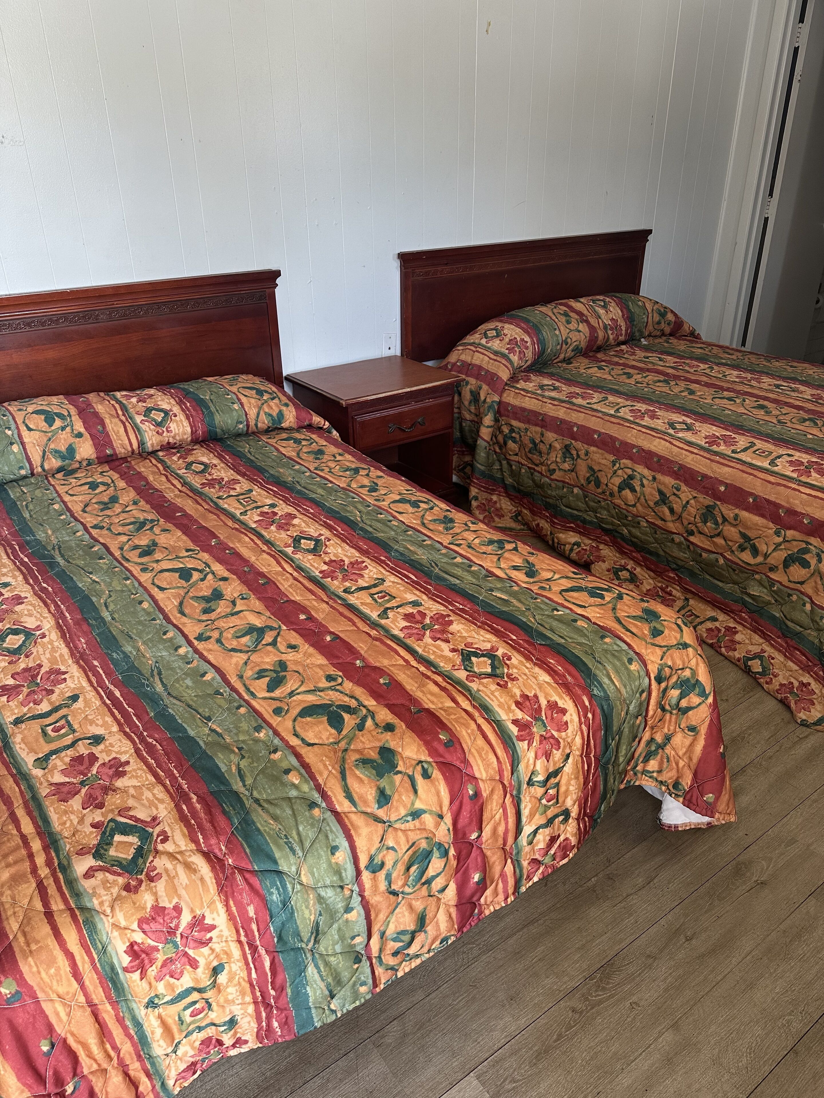 A room with two wooden beds, each with a patterned bedspread and matching pillows. There is a wooden nightstand between the beds.