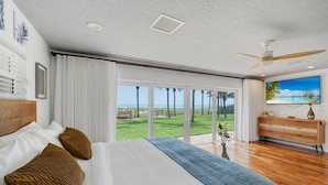 A bedroom with a large bed, a view of the outdoors, a ceiling fan, and a TV on a wooden cabinet. - Dream Beach Home Vacation - Direct Beach Access & Exquisite Waterfront Views (Cocoa Beach)
