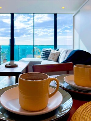 Ein modernes Zimmer mit großem Fenster und Blick auf eine Stadtlandschaft. Ein Tisch mit gelber Tasse und Untertasse sowie ein Sessel mit Kissen.