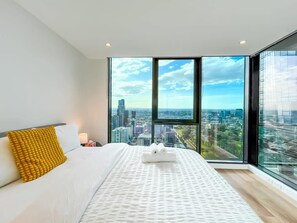 Ein modernes Hotelzimmer mit einem großen Bett, einem gelben Kissen und Blick auf die Stadt durch raumhohe Fenster.