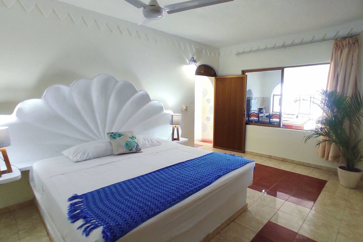 A hotel room with a large bed, a ceiling fan, a nightstand with a lamp, a window with curtains, and a potted plant.