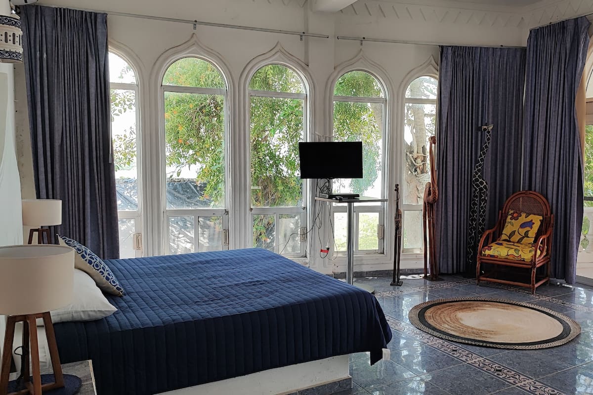 A bedroom with a large bed, a television on a stand, a chair, and large windows with curtains.