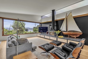 A modern living room with a large window, a sailboat model, a fireplace, and a TV. - 24 Hill Street - Pet friendly (Merimbula)