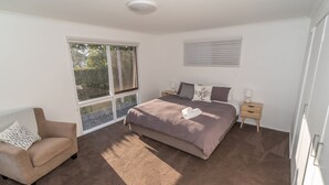 A bedroom with a large window, a bed with a beige blanket, a bedside table with a lamp, and a chair with a patterned cushion. - Lochview - Pet Friendly Merimbula (Merimbula)