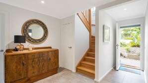 Un couloir avec un escalier en bois, une armoire en bois, un miroir doté d’un cadre orné et une porte donnant sur un espace extérieur agrémenté de plantes.