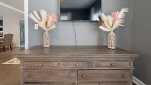 A wooden dresser with two vases of dried flowers, a flat-screen TV mounted on the wall, and a dining area visible in the background. - Walk to Downtown~3 King Beds~Private Office (Raleigh)