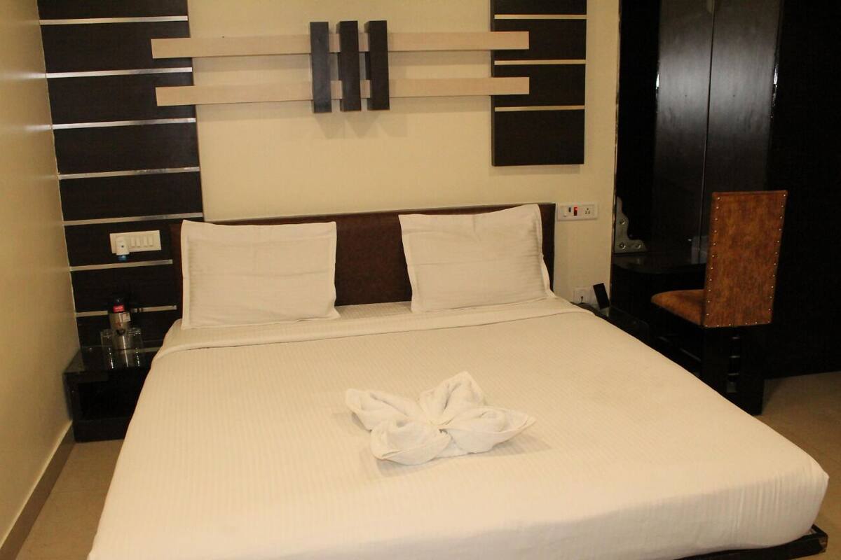 Habitación de hotel con una cama grande, dos almohadas, una toalla doblada sobre la cama, una mesita de noche con una botella y una silla.