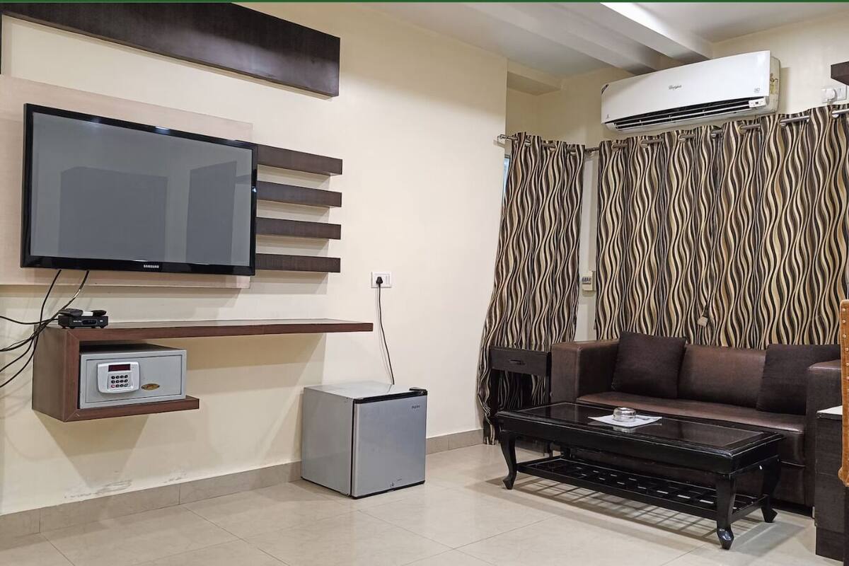 Una sala de estar moderna con un televisor de pantalla plana montado en un estante de madera, un refrigerador pequeño, un sofá y una cortina estampada.
