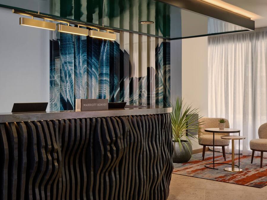Un moderno mostrador de recepción con un diseño ondulado único, una laptop y un letrero que dice "MARRIOTT BONVVEY". Se ven dos sillas y una pequeña mesa al fondo, junto a un ventanal con cortinas.