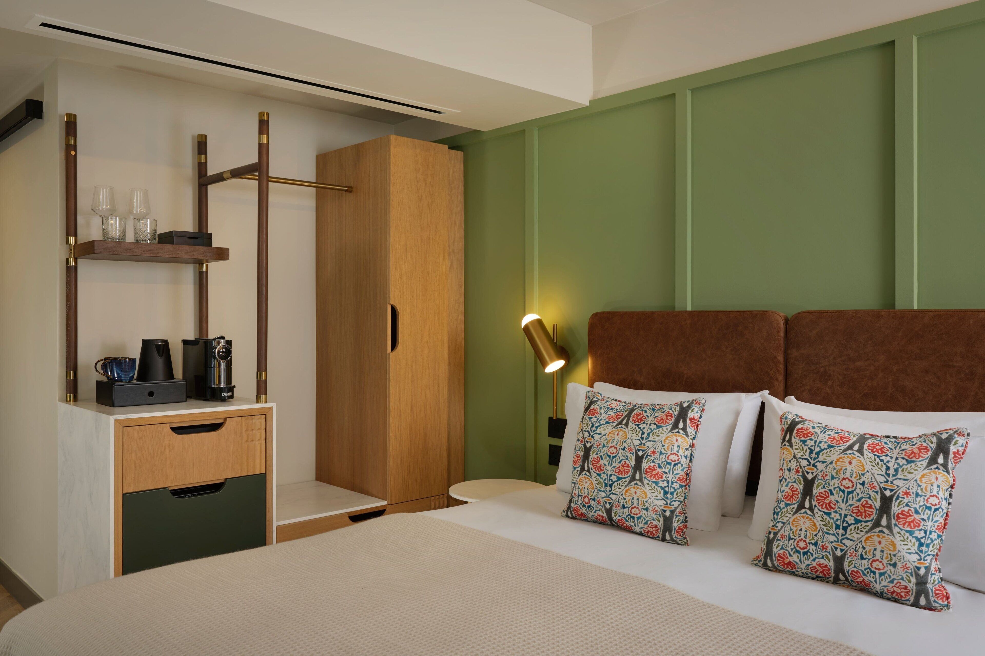 Habitación de hotel impecable con cama, armario de madera, mesita de noche con lámpara y una pequeña estantería con vasos y cafetera.