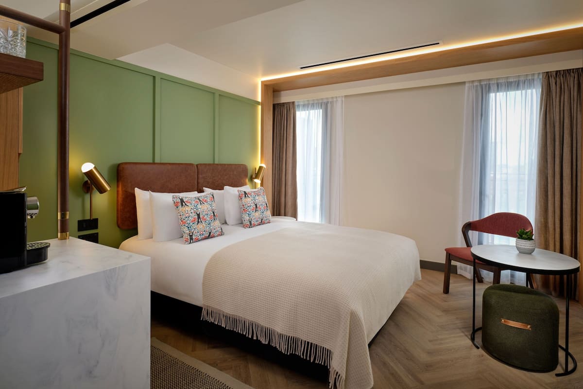 A hotel room with a large bed, a desk with a lamp, a chair, and a small table with a plant.