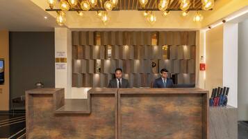 A reception desk with two staff members, a wooden panel wall, and a ceiling with hanging light fixtures.
