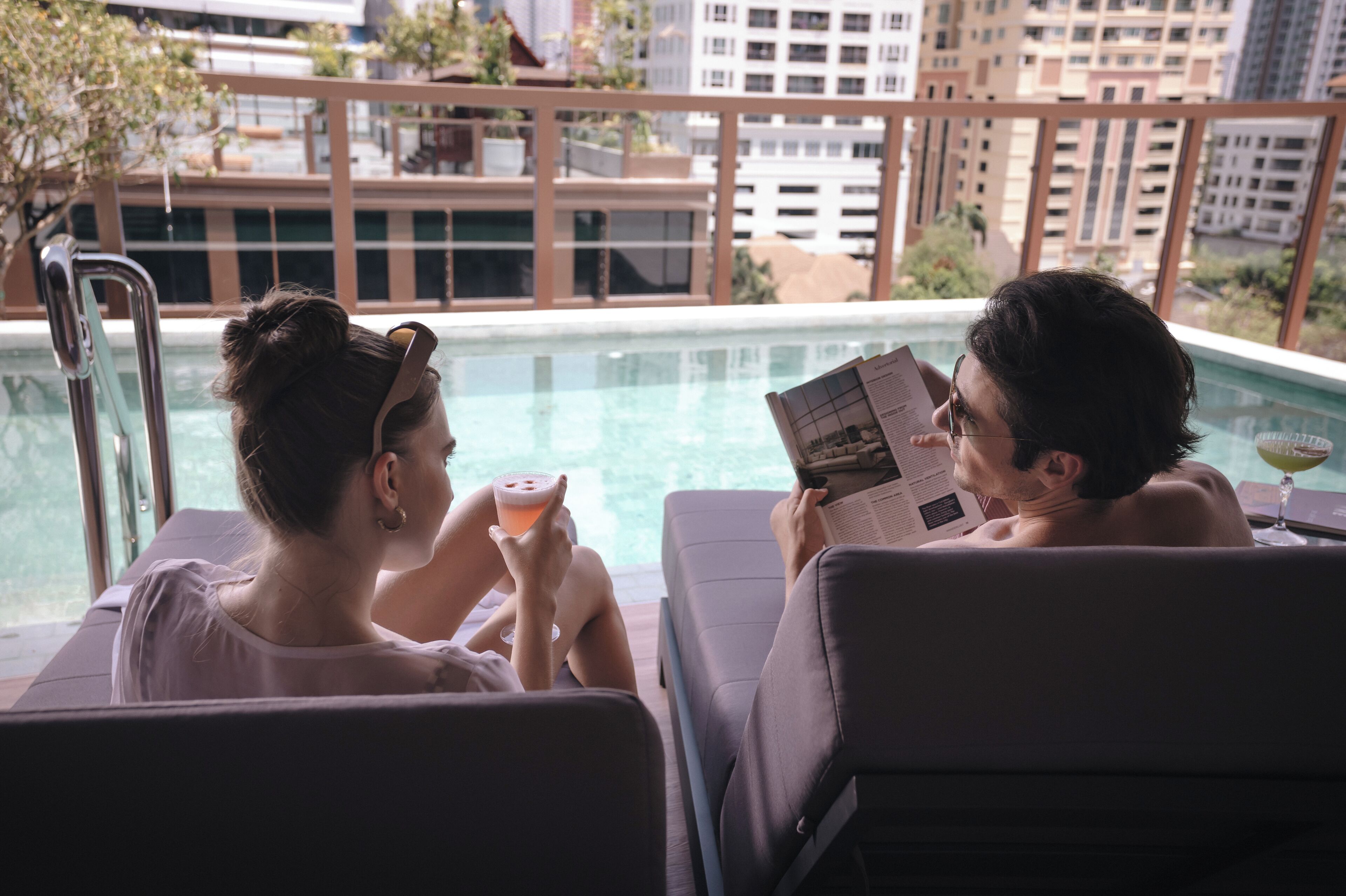 Two people relaxing by a poolside lounge chair, reading a book and holding a drink.