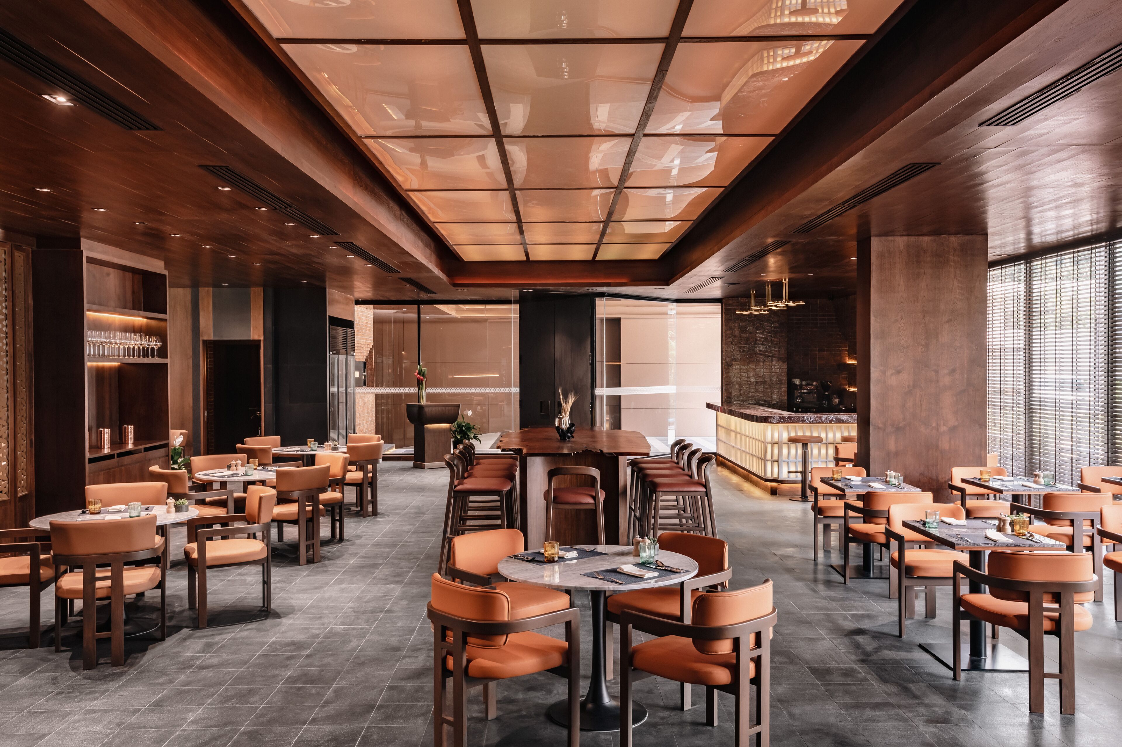 A spacious dining area with round tables and orange chairs, featuring a modern ceiling design and a bar area in the background.