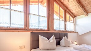 Un dormitorio con cama, almohadas, una mesita de noche con lámpara y una ventana con cortinas.
