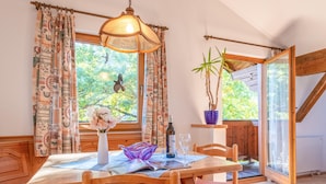 Un comedor con una mesa de madera, sillas, un jarrón con flores, una botella de vino y una copa. Se ve un ventanal con cortinas y vistas a la vegetación exterior.