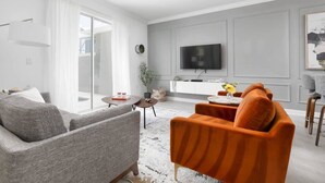 Ett modernt vardagsrum med en grå soffa, en orange fåtölj, en platt-TV och en matplats med ett litet bord och stolar.