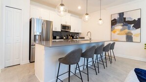 A modern kitchen with a central island, bar stools, stainless steel appliances, pendant lighting, and a large abstract painting on the wall. - Chic Modern, Themed Bedrooms, Private Pool (Kissimmee)