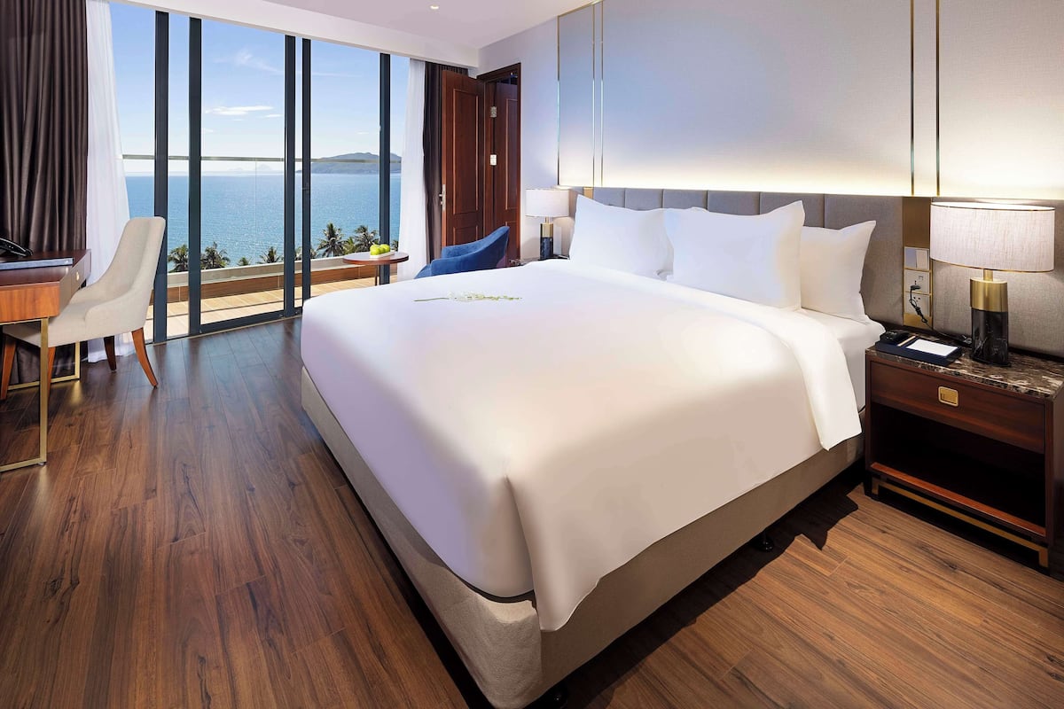 A modern hotel room with a large bed, a desk, and a chair. The room has a view of the sea.