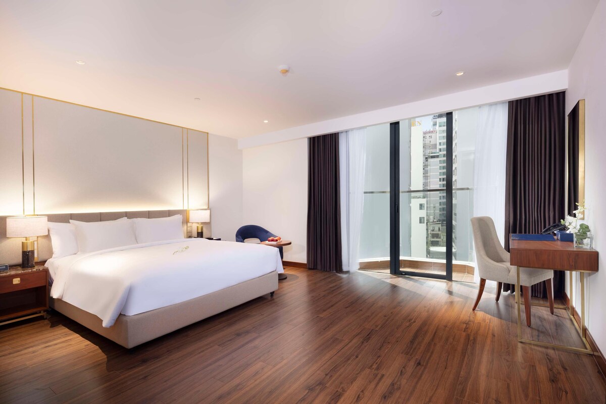 A hotel room with a large bed, a desk, a chair, and a view of the city through a glass door.