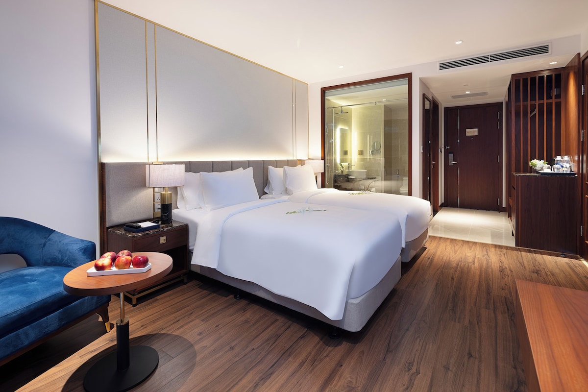 A modern hotel room with a large bed, a bedside table with a lamp, a small round table with fruit, and a blue sofa.