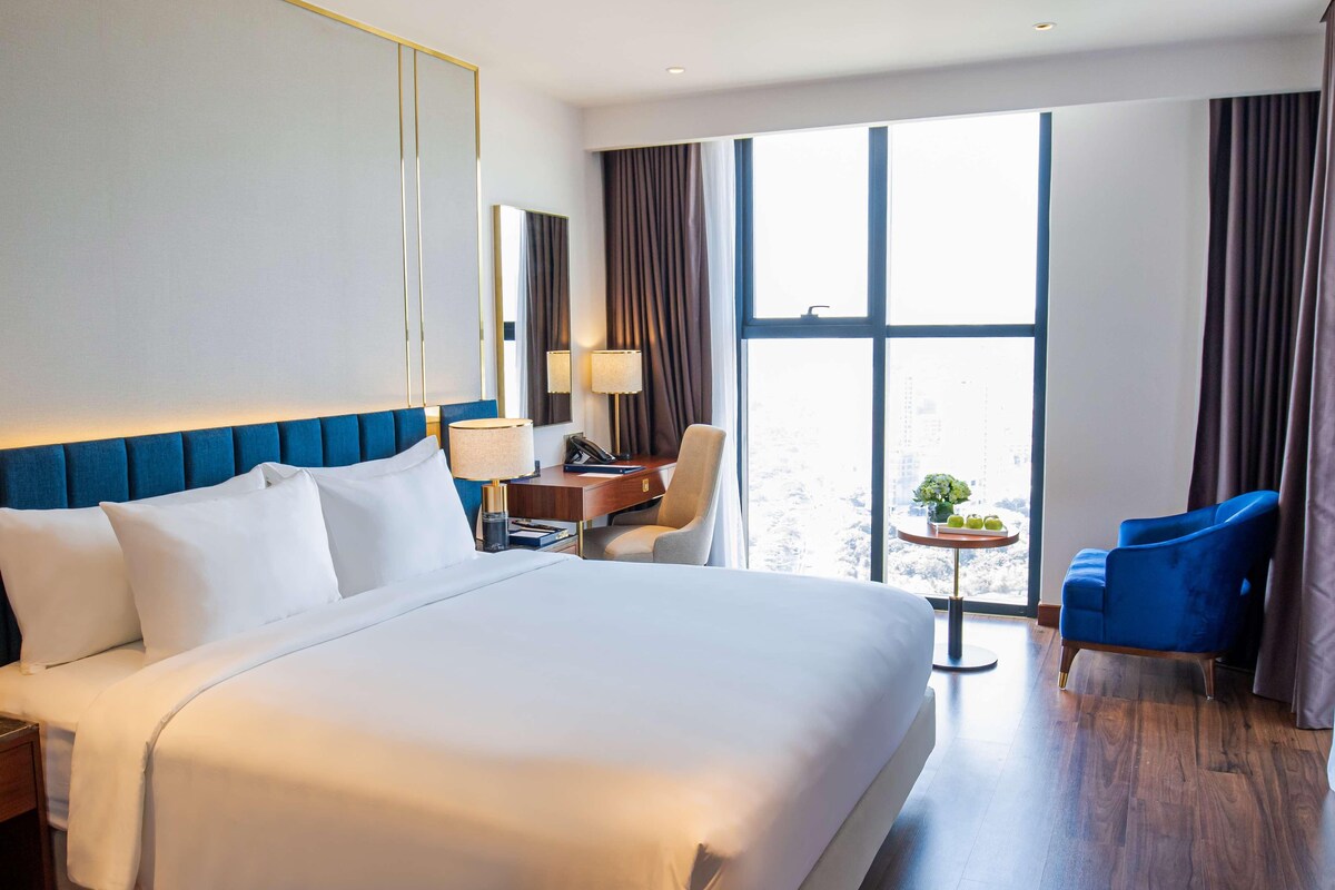A hotel room with a large bed, a desk with a lamp, a chair, and a small table with a plant.