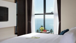 A hotel room with a large bed, a flat-screen TV mounted on the wall, a view of the ocean through a large window, and a small table with a vase of flowers and a fruit bowl. - Best Western Premier Marvella Nha Trang (Nha Trang)