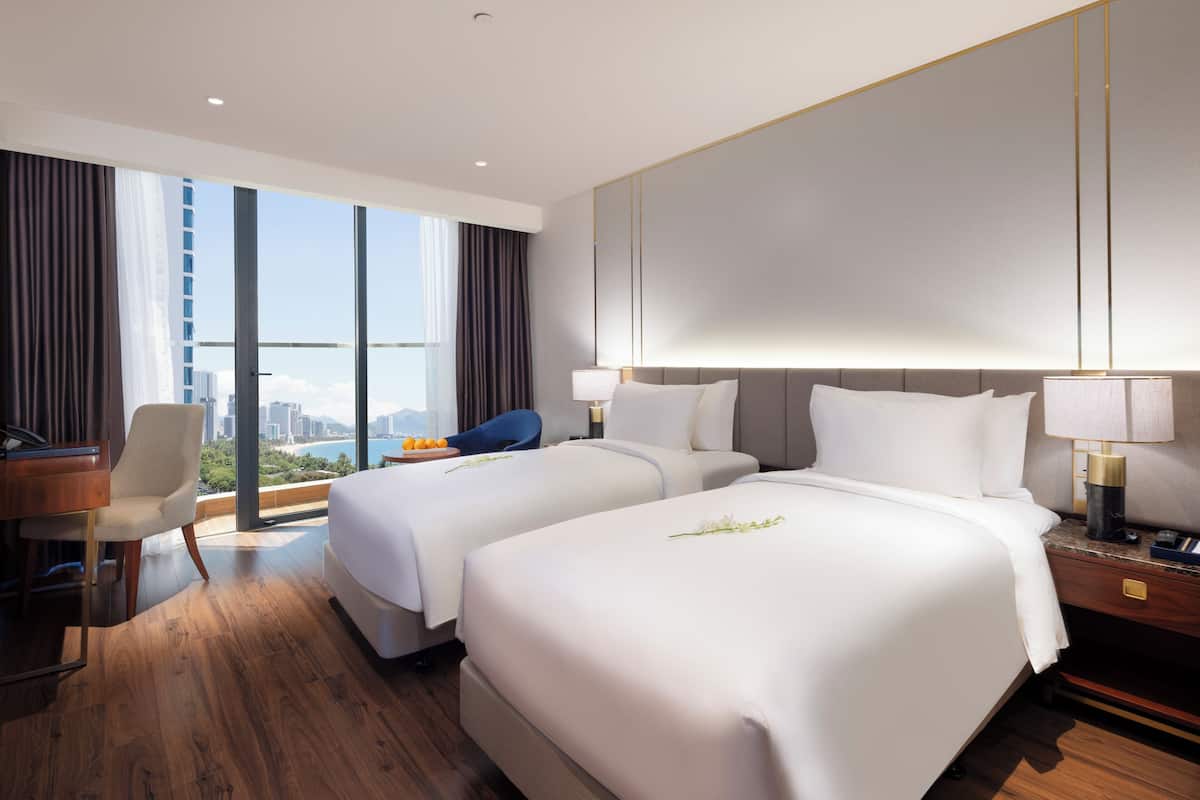 A hotel room with two beds, a desk, a chair, and a view of the city.
