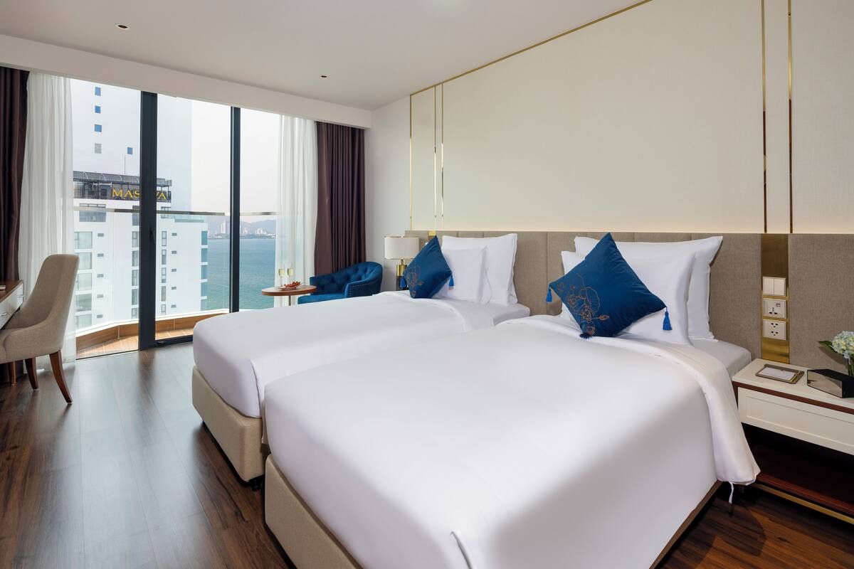 A large bed with white linens, a bedside table with a lamp, and a view of a cityscape through large windows.