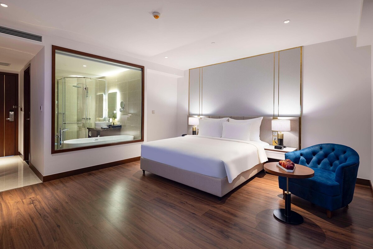 A modern hotel room with a large bed, a sitting area with a blue chair, a small round table, and a large mirror reflecting the bathroom.