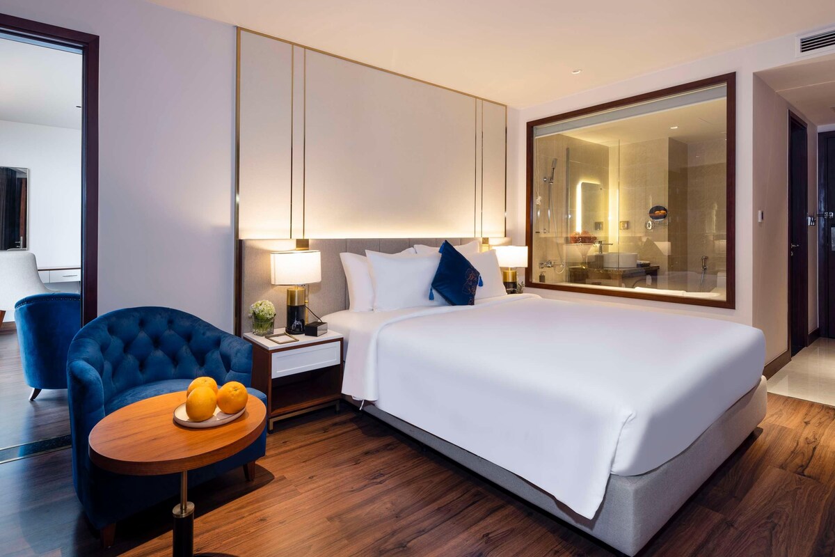 A hotel room with a large bed, a bedside table with a lamp, a blue armchair, a small round table with oranges, and a large mirror.