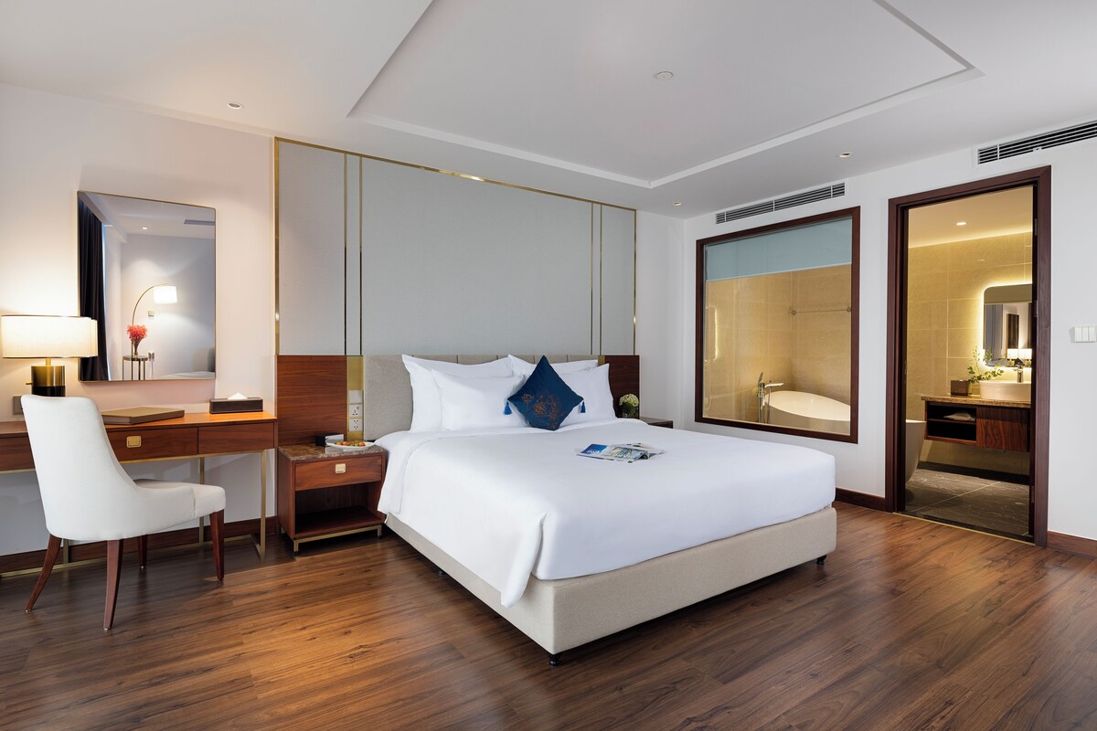 A modern hotel room with a large bed, a desk, a chair, and a bathroom visible through an open door.
