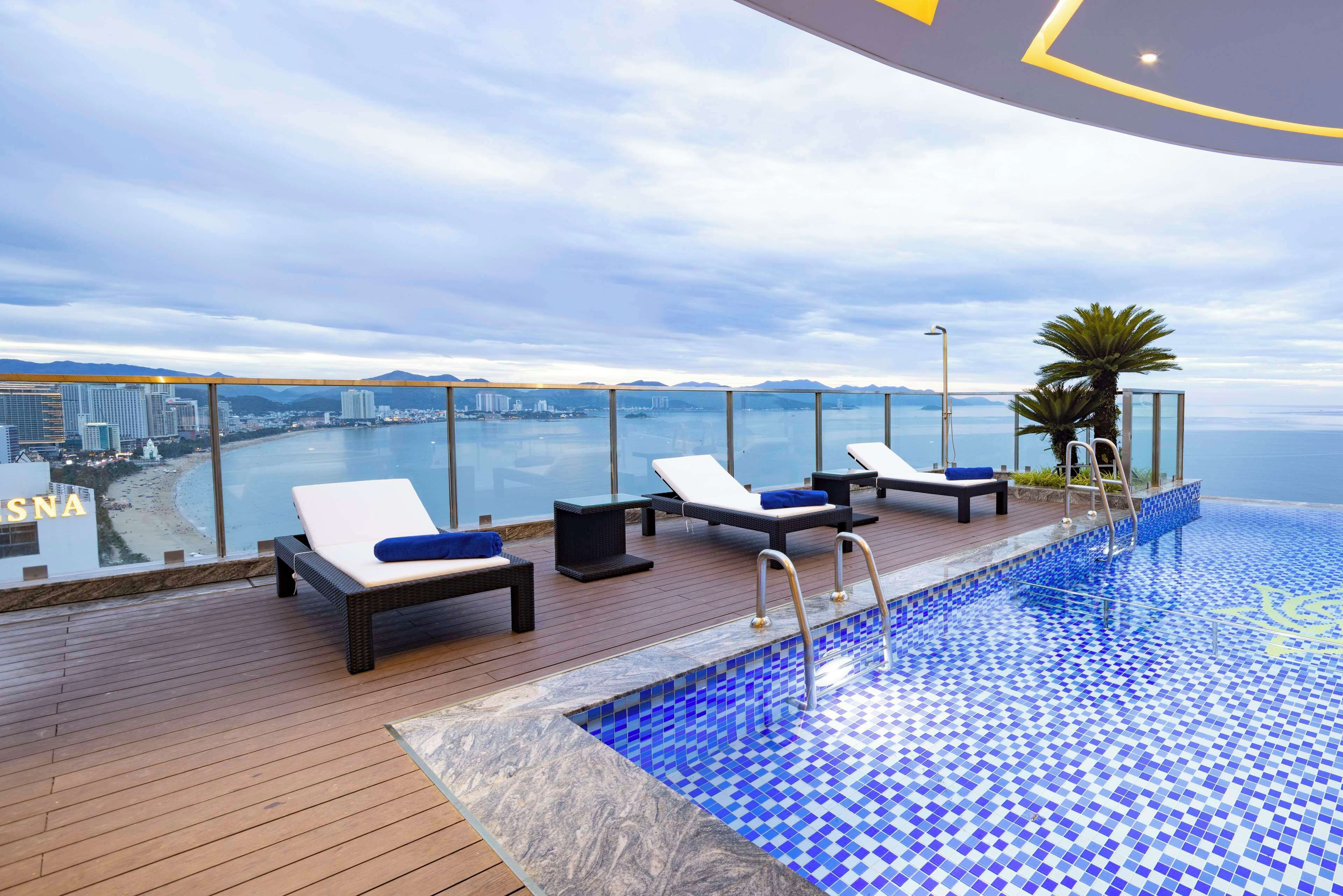 A rooftop infinity pool with lounge chairs and a wooden deck overlooking a cityscape and beach.