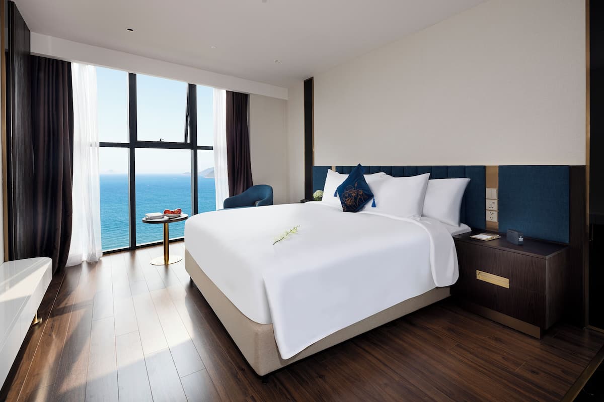 A modern hotel room with a large bed, a bedside table, a chair, and a view of the sea through large windows.