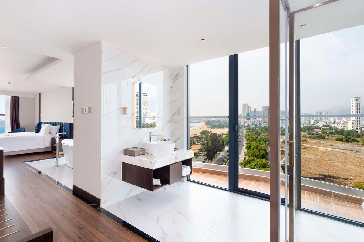 A modern hotel room with a large bed, a desk, and a bathroom with a large mirror. The room has a city view.