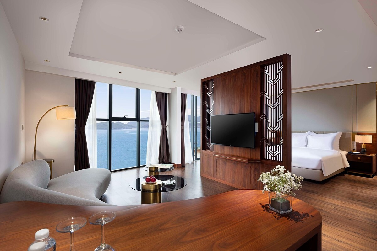 A modern hotel room with a large window offering a sea view, a flat-screen TV mounted on a wooden panel, a comfortable sofa, a wooden coffee table with a vase of flowers, and a neatly made bed.