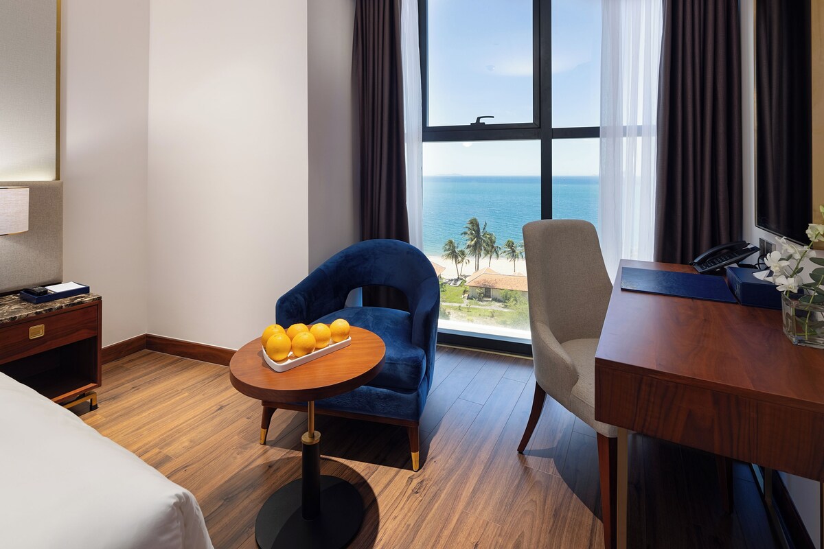 A hotel room with a bed, a desk with a telephone, a chair, a small table with fruit, and a view of the ocean through large windows.