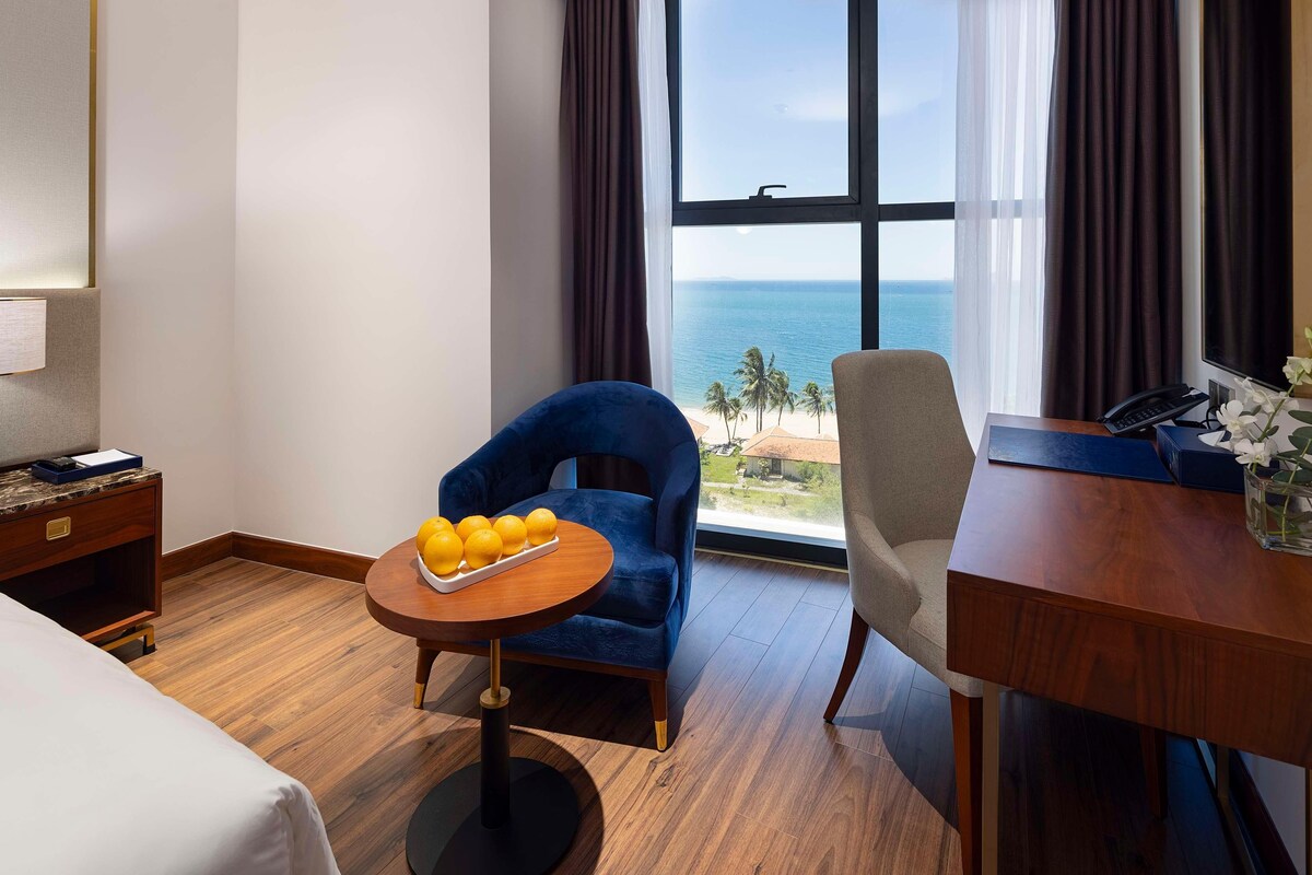 A hotel room with a bed, a desk, a chair, a small table with fruit, and a large window with a view of the beach and ocean.