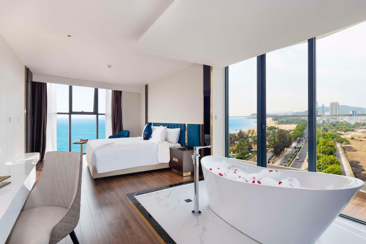 A modern hotel room with a large bed, a sitting area, a bathtub with rose petals, a large window with a city view, and a minibar.