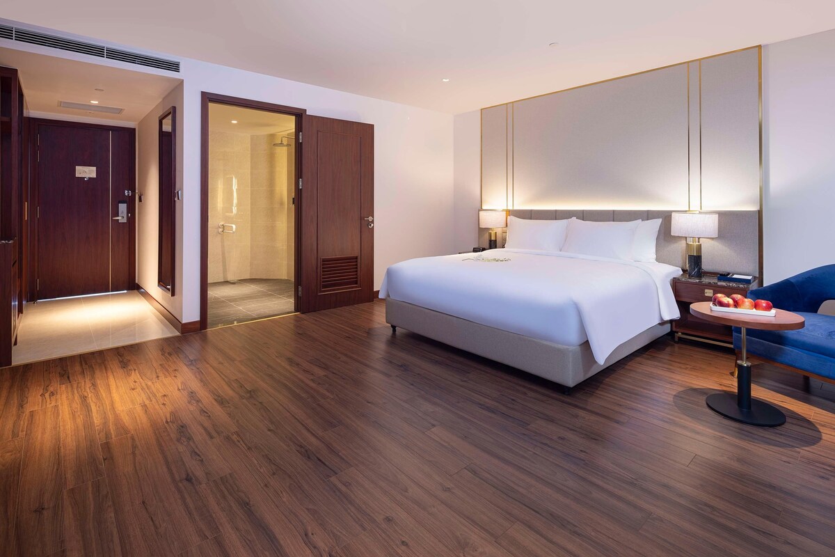 A hotel room with a large bed, bedside tables, a small round table with fruit, and a blue sofa.