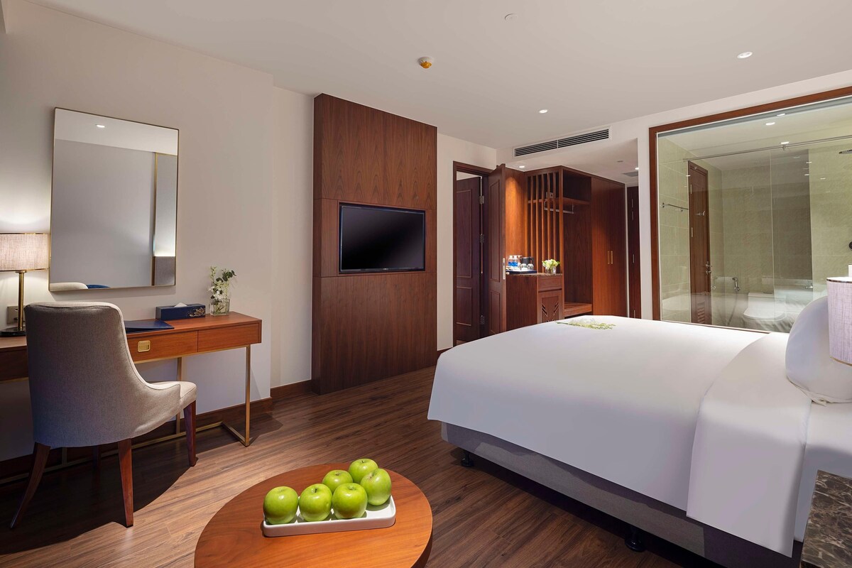 A hotel room with a bed, a desk, a chair, a TV, and a bathroom visible through a glass door.